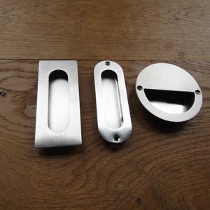 Op de afbeelding: Drie verzonken handgrepen van geborsteld metaal in rechthoekige, ovale en ronde vormen, vergezeld van twee kleine metalen schroeven. Elke handgreep heeft een verzonken greepgedeelte voor schuifdeuren of lades met een gladde afwerking.