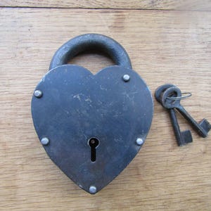 Peut inclure: Un cadenas en métal gris foncé en forme de cœur avec une anse arrondie sur le dessus. Le cadenas présente une serrure centrale et cinq rivets visibles sur son pourtour. Deux clés en métal assorties sont fixées à un petit anneau à côté.