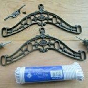 May include: Two dark bronze-coloured metal clothes dryer brackets with ornate scrollwork, two metal pulleys with threaded bolts, a small metal cleat with a screw, and a roll of white clothesline rope wrapped in blue and white plastic packaging.