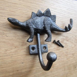 Könnte beinhalten: Ein dunkelgrauer Wandhaken aus Gusseisen in Form eines Stegosaurus-Dinosauriers. Der Haken hat eine texturierte, metallische Oberfläche mit einem geschwungenen unteren Arm, der in einem abgerundeten Knopf endet. Zwei passende Metallschrauben liegen bei.