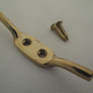 May include: A polished brass boat cleat featuring a central mounting plate with two countersunk screw holes, accompanied by two matching brass wood screws. The hardware has a smooth, reflective metallic finish and a classic dual-horn design.