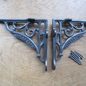 May include: Two dark gray metal shelf brackets with an ornate scrollwork design and the text SINGER CORP embossed on the diagonal support bar. The brackets feature mounting holes at the top and side, accompanied by a small pile of metal screws.
