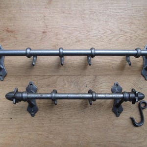 May include: Two dark gray metal wall-mounted racks with horizontal bars and multiple hooks. The racks feature decorative end caps and mounting brackets. A separate S-shaped hook and a small plastic bag containing black screws are included.