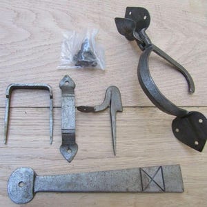Op de afbeelding: Een set donkergrijze metalen hardwarecomponenten, waaronder een grote deurklink met hartvormige montageplaten, een U-vormige kram, een platte rechthoekige grendel, een puntige haak, een lange taps toelopende heng en een klein zakje met schroeven.