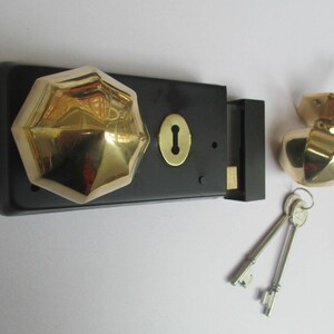 K&ouml;nnte beinhalten: Ein schwarzer, rechteckiger T&uuml;rschlossmechanismus mit einem polierten, goldfarbenen, achteckigen Knauf und einer passenden goldfarbenen Schl&uuml;ssellochplatte. Enthalten ist ein separater, achteckiger Knauf mit Metallstift und zwei silberne Schl&uuml;ssel.