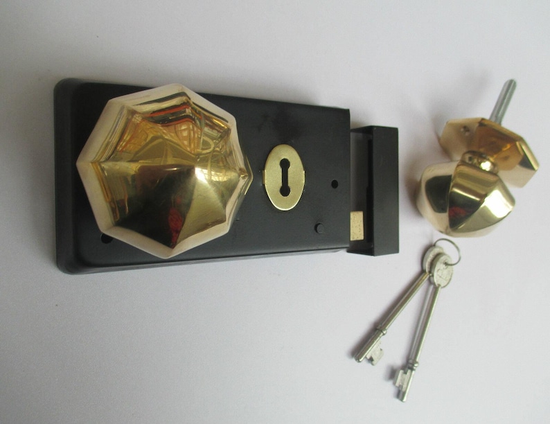 K&ouml;nnte beinhalten: Ein schwarzer, rechteckiger T&uuml;rschlossmechanismus mit einem polierten, goldfarbenen, achteckigen Knauf und einer passenden goldfarbenen Schl&uuml;ssellochplatte. Enthalten ist ein separater, achteckiger Knauf mit Metallstift und zwei silberne Schl&uuml;ssel.