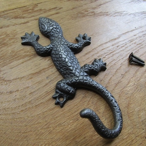 Puede incluir: Un gancho de pared de metal gris oscuro texturizado con forma de lagarto y cola curva. La figura del lagarto presenta escamas detalladas en todo el cuerpo y cuatro patas extendidas. Dos pequeños tornillos de metal oscuro se sitúan a la derecha.