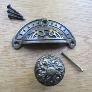 Puede incluir: Un tirador de cajón de metal semicircular con un patrón en relieve y dos orificios para tornillos, acompañado de un pomo de armario redondo de metal con un diseño floral texturizado, tres tornillos negros pequeños y un perno de montaje plateado largo.