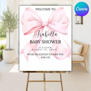 May include: A baby shower sign with a large pink bow graphic. The sign reads "Welcome to Isabella Baby Shower" with the date and time. The sign is displayed on a gold easel.