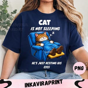 May include: A navy blue t-shirt featuring a graphic of a brown tabby cat resting in a blue recliner chair under a blue blanket. The text reads CAT IS NOT SLEEPING HE'S JUST RESTING HIS EYES with Zz symbols near the cat's head, printed in white.