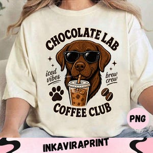 May include: A cream-coloured t-shirt featuring a graphic of a brown Labrador dog wearing black sunglasses and drinking iced coffee. The text reads Chocolate Lab Coffee Club, iced vibes, and brew crew, accompanied by paw print and coffee bean icons.