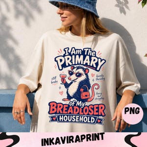 Pode incluir: Uma t-shirt bege com o gráfico de um gambá usando óculos de sol, segurando uma taça de vinho e uma bolsa. O texto diz: I Am The PRIMARY of My BREADLOSER HOUSEHOLD, com as frases: still broke, barely thriving, fancy but struggling.