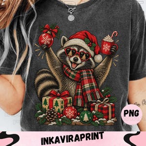 Puede incluir: Una camiseta gris carbón con el gráfico de un mapache que lleva un gorro de Papá Noel rojo, gafas en forma de corazón y una bufanda de cuadros. El mapache sostiene un adorno rojo y una taza, rodeado de regalos, piñas y ramas de pino.