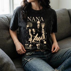 Pu&ograve; includere: T-shirt grigio scuro con una grafica della band "NANA black stones". Il design include illustrazioni dei membri della band e testo. La maglietta &egrave; indossata da una persona in jeans blu.
