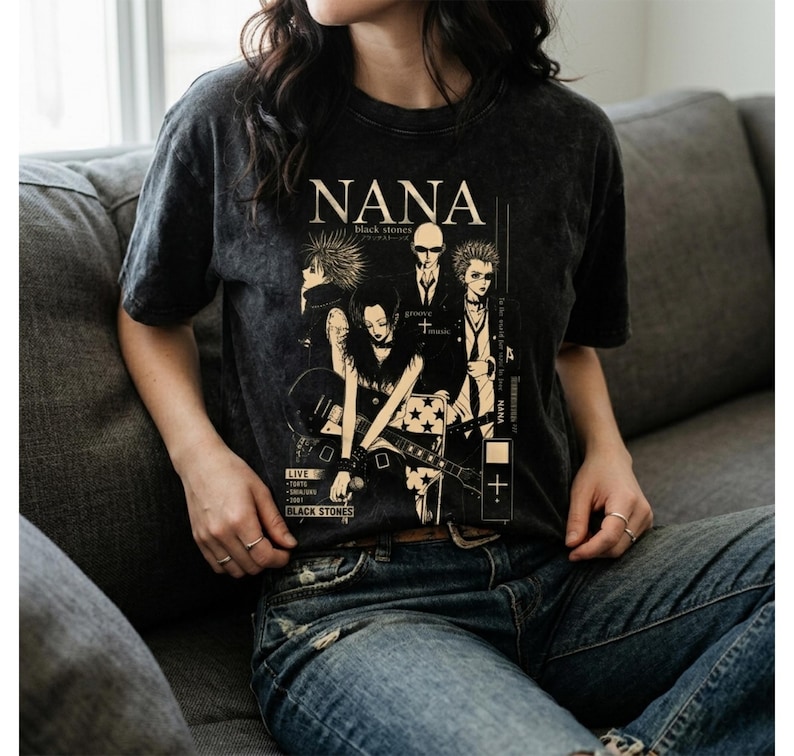 Pu&ograve; includere: T-shirt grigio scuro con una grafica della band "NANA black stones". Il design include illustrazioni dei membri della band e testo. La maglietta &egrave; indossata da una persona in jeans blu.
