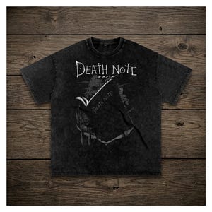 Può includere: Una maglietta nera con le parole "Death Note" in bianco, insieme a caratteri giapponesi. La maglietta presenta un design grafico della serie anime. La maglietta è esposta su una superficie di legno.