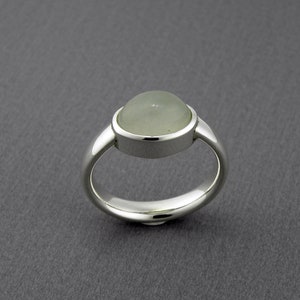May include: A silver ring with a large, round, pale green gemstone. The gemstone is set in a bezel setting, and the band is smooth and polished. The ring is displayed on a gray background.