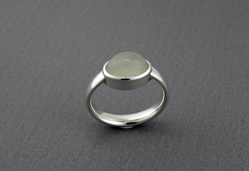 May include: A silver ring with a large, round, pale green gemstone. The gemstone is set in a bezel setting, and the band is smooth and polished. The ring is displayed on a gray background.