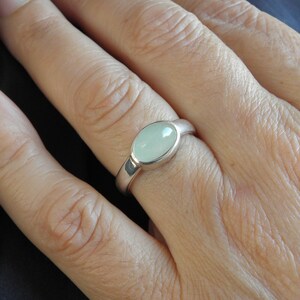 May include: A silver ring with a light green oval stone. The ring is set in a simple, polished band. The stone has a smooth, polished surface. The ring is shown on a person's finger.