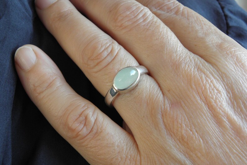 May include: A silver ring with a light green oval stone. The ring is set in a simple, polished band. The stone has a smooth, polished surface. The ring is shown on a person's finger.