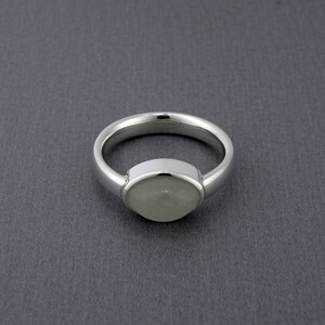 May include: A silver ring with a light green, oval-shaped stone set in a bezel setting. The band is smooth and polished, and the ring is displayed against a dark gray background. The ring is a piece of jewelry.