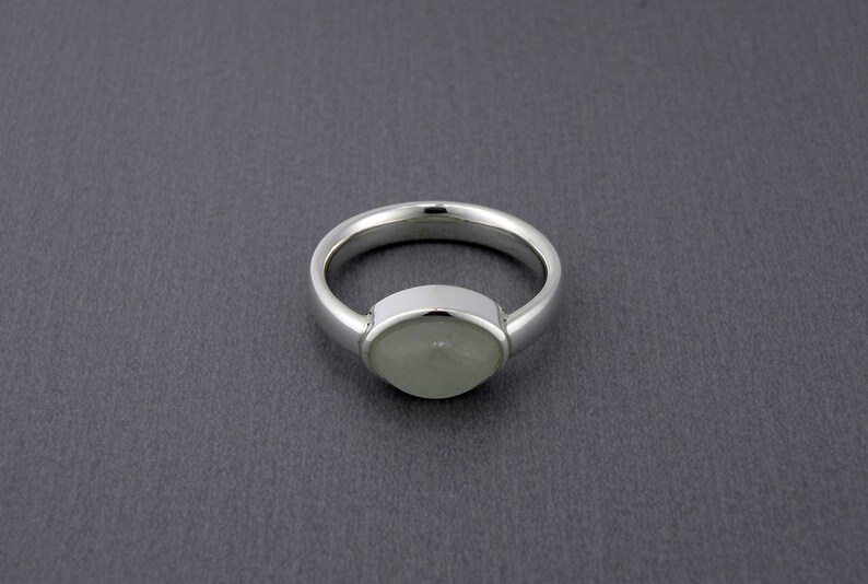 May include: A silver ring with a light green, oval-shaped stone set in a bezel setting. The band is smooth and polished, and the ring is displayed against a dark gray background. The ring is a piece of jewelry.