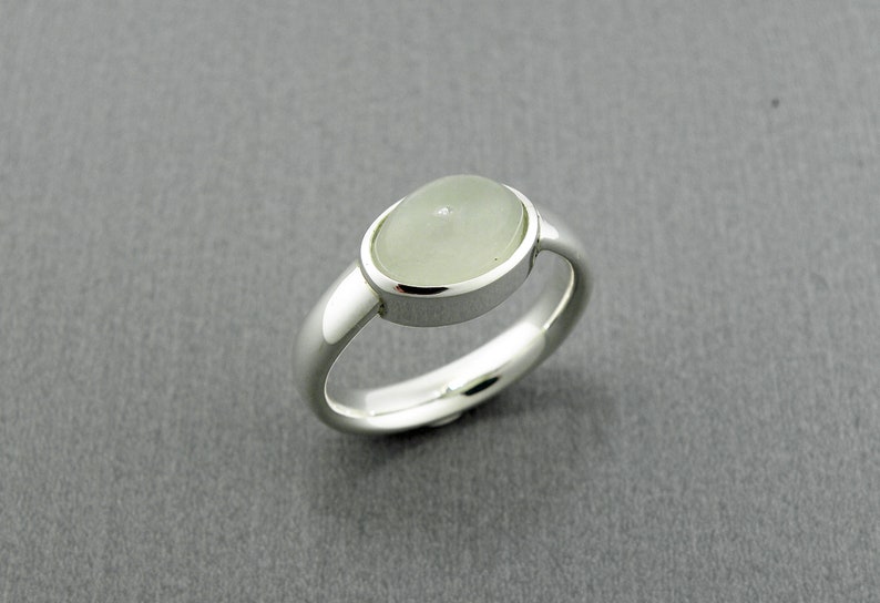 May include: A silver ring with a light green, oval-shaped stone. The ring has a smooth, polished band and a bezel setting that holds the stone securely. The ring is displayed on a gray background.