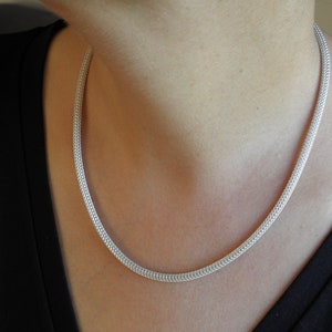 Può includere: Una collana a catena d'argento con un design delicato e intrecciato. La catena è indossata da una persona con una camicia nera.