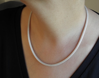 Könnte beinhalten: Eine silberne Kettenkette mit einem zarten, gewebten Design. Die Kette wird von einer Person mit einem schwarzen Shirt getragen.