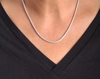 Könnte beinhalten: Eine silberne Halskette mit geflochtenem Design wird um den Hals getragen. Die Halskette wird vor einem schwarzen V-Ausschnitt-Shirt präsentiert. Die Kette hat eine helle, glänzende silberne Farbe.