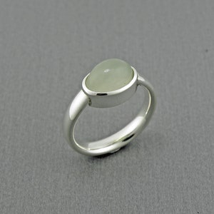 May include: A silver ring featuring a pale green, oval-shaped gemstone. The gemstone is set in a bezel setting, and the ring has a polished, rounded band. The ring is a piece of jewelry.