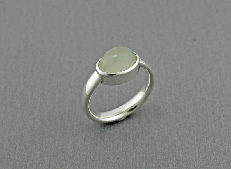May include: A silver ring featuring a pale green, oval-shaped gemstone. The gemstone is set in a bezel setting, and the ring has a polished, rounded band. The ring is a piece of jewelry.