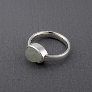 May include: A silver ring with a pear-shaped, pale green gemstone. The ring has a smooth, polished band and a bezel setting that holds the stone securely. The ring is displayed on a gray surface.