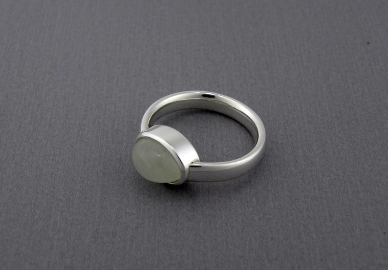 May include: A silver ring with a pear-shaped, pale green gemstone. The ring has a smooth, polished band and a bezel setting that holds the stone securely. The ring is displayed on a gray surface.