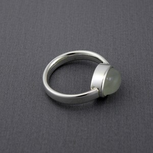 May include: A silver ring with a rounded band and a bezel-set, light green, oval-shaped gemstone. The ring is set against a dark gray background, showcasing the polished metal and the translucent quality of the stone.