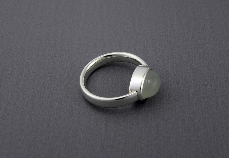 May include: A silver ring with a rounded band and a bezel-set, light green, oval-shaped gemstone. The ring is set against a dark gray background, showcasing the polished metal and the translucent quality of the stone.