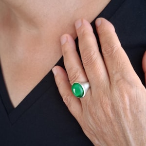 Op de afbeelding: Een zilveren ring met een gladde, ovaalvormige, levendig groene cabochon edelsteen gezet in een gepolijste metalen band. De ring heeft een substantieel, afgerond profiel met een strakke metalen afwerking die contrasteert met de groene steen.
