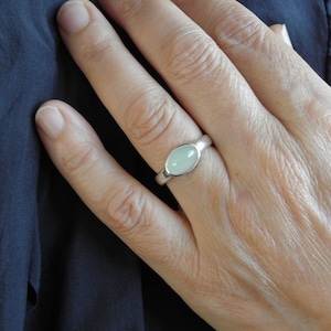 May include: A silver ring with an oval, pale green gemstone. The ring is set in a simple, polished band. The hand wearing the ring is resting on a dark blue fabric.
