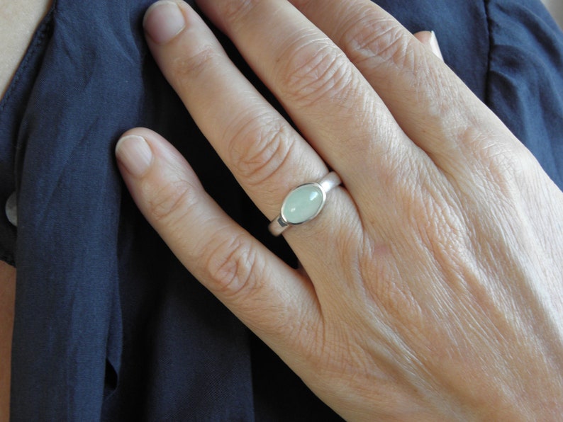 May include: A silver ring with an oval, pale green gemstone. The ring is set in a simple, polished band. The hand wearing the ring is resting on a dark blue fabric.
