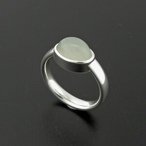 May include: A silver ring with a light green, oval-shaped gemstone. The gemstone is set in a bezel setting, and the band is smooth and polished. The ring is displayed on a dark surface, highlighting its reflective qualities.