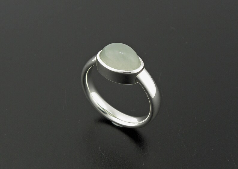May include: A silver ring with a light green, oval-shaped gemstone. The gemstone is set in a bezel setting, and the band is smooth and polished. The ring is displayed on a dark surface, highlighting its reflective qualities.