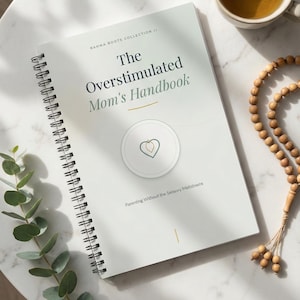 May include: A handbook titled "The Overstimulated Mom's Handbook" with a spiral binding, featuring a heart-shaped logo. The book is on a white marble surface, with a cup of tea, wooden beads, and greenery. The subtitle reads "Parenting Without the Sensory Meltdowns."