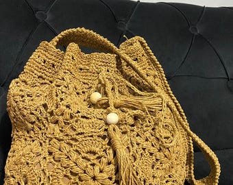 Könnte beinhalten: Handgefertigte Häkeltasche in warmem Goldbraun. Die Tasche hat einen Kordelzugverschluss, einen Schulterriemen und dekorative Quasten mit Holzperlen. Das florale Muster verleiht einen Hauch von Bohème-Stil.