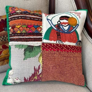 May include: Square patchwork pillow with a green border. The pillow features various fabric patterns, including floral, embroidered, and textured designs in shades of orange, red, green, and white. An embroidered figure is on one panel.