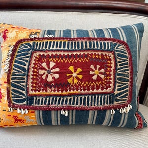 May include: A blue and yellow patchwork pillow with a red embroidered center featuring three white flowers. The pillow is decorated with white shell-like embellishments.