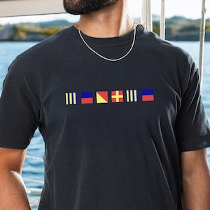 May include: Dark navy blue t-shirt featuring a nautical flag design in yellow, red, blue, and white. The shirt has a crew neck and short sleeves. The design is centered on the chest. The background shows a body of water.