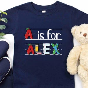 May include: A navy blue short-sleeved t-shirt featuring a graphic print with the text "A is for ALEX". The letters are stylized as school-themed objects including a red apple, a yellow pencil, lined paper, and a blue and green globe.
