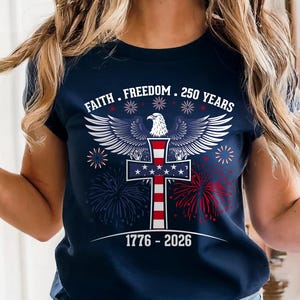 May include: Navy blue t-shirt with a patriotic design. The shirt features a cross with the American flag, an eagle, and the text "Faith, Freedom, 250 Years" and "1776-2026". Red and blue fireworks are also included in the design.