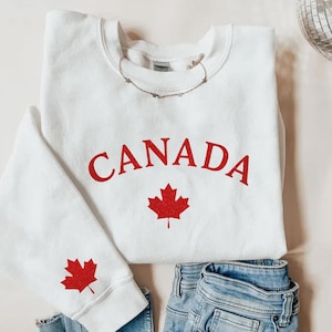 May include: White sweatshirt with the word "CANADA" in red arched lettering and a red maple leaf graphic. A red maple leaf is also on the left sleeve. The sweatshirt is paired with blue jeans.