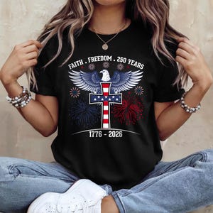 May include: Black t-shirt with a patriotic design featuring an eagle, cross, and fireworks. The text reads "Faith. Freedom. 250 Years" above the cross, and "1776-2026" below. The design incorporates red, white, and blue colors.